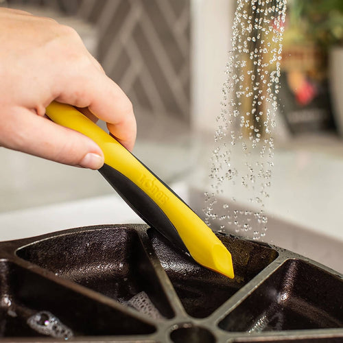 Hand using a yellow and black dual-head brush to clean grooves of a cast iron pan under running water, ideal for Kitchen & Food, Cookware, Cast Iron, Best Sellers, and Promo Eligible.