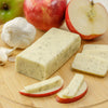 Handcrafted block of shelf-stable cheese with herb flecks, sliced and paired with fresh apple wedges and garlic on a wooden cutting board, ideal for Kitchen & Food and Pantry collections.