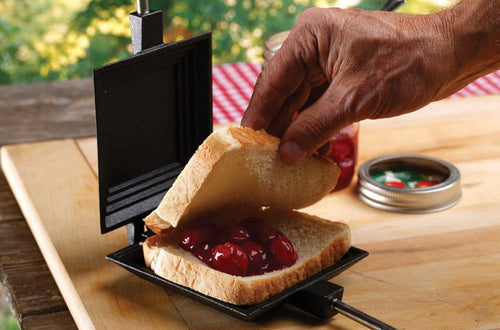 Cast iron square pie iron with metal handle and wood grips, shown with bread and cherry pie filling being prepared, ideal for camping and included in Farm, Garden & Tool, Camping, Best Sellers, and Promo Eligible collections.