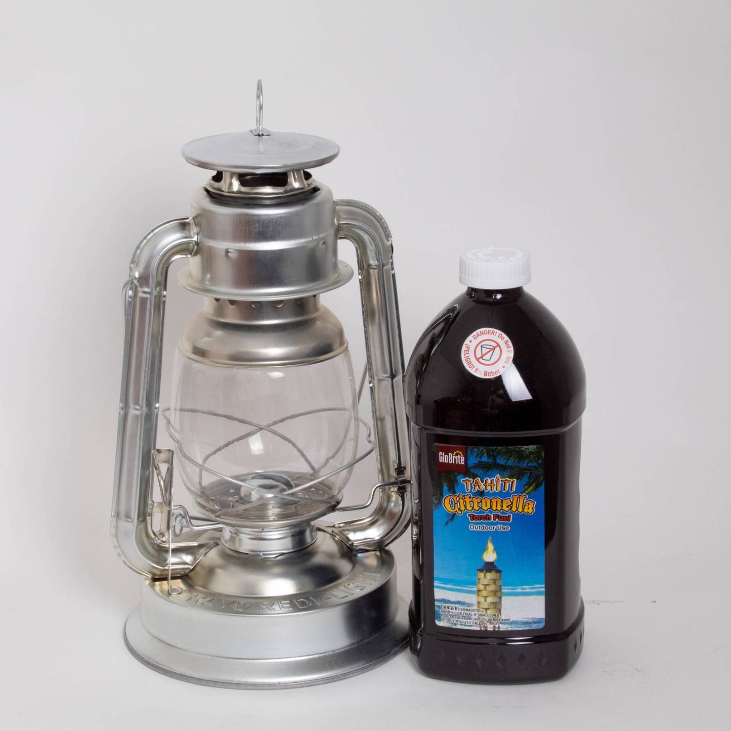 Silver metal oil lantern with clear glass globe standing next to a large brown plastic bottle of citronella oil, suitable for outdoor use in farm, garden, camping, pest control, and emergency supplies collections.