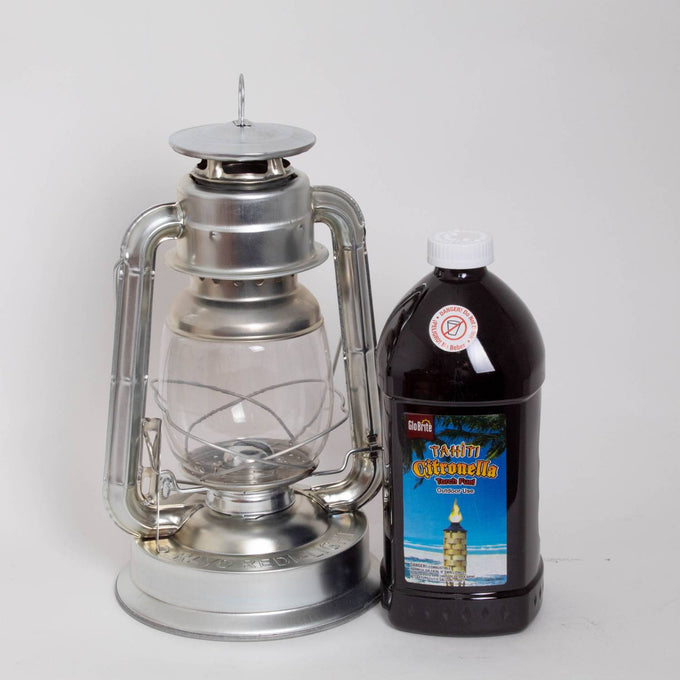Silver metal oil lantern with clear glass globe standing next to a large brown plastic bottle of citronella oil, suitable for outdoor use in farm, garden, camping, pest control, and emergency supplies collections.