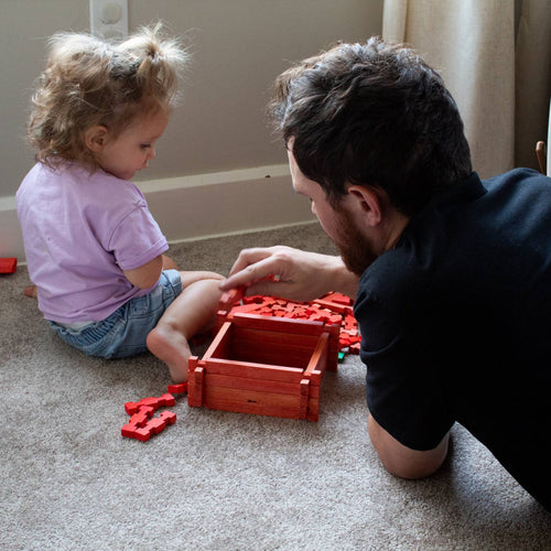 Father and young child playing together indoors with handcrafted wooden log pieces in red, encouraging creative building and imaginative play with a compact toy set made in USA.