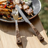 Fork and serrated knife with engraved wooden handles resting on a wooden surface beside a metal plate of grilled food, part of a camping cutlery set featured in Farm, Garden & Tool, Kitchen & Food, and Camping collections.
