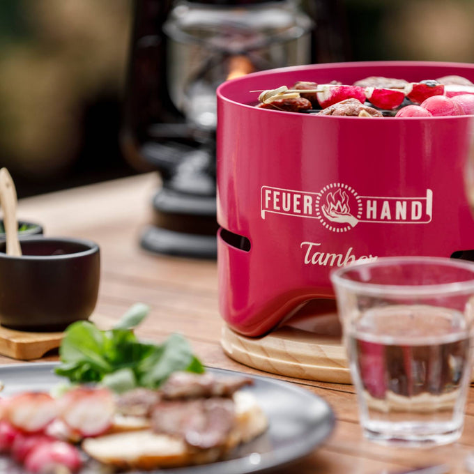 Bright red Feuerhand Tamber tabletop grill with vibrant skewers cooking over low heat, set on a wooden trivet on an outdoor table, featured in Farm, Garden & Tool and Grilling collections.