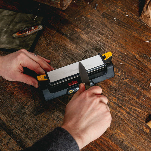 Hands sharpening a knife using a compact benchstone sharpener with coarse and fine abrasive surfaces, on a rustic wooden workbench, suitable for farm, garden, and kitchen use.