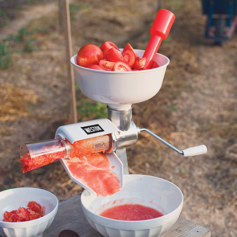 Manual tomato press with white hopper filled with Roma tomatoes, featuring a red plunger and metal crank, straining fresh tomato sauce into white bowls outdoors, part of Kitchen & Food, Food Prep, Food Mills, and Best Sellers collections.