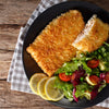 Crispy golden breaded fish fillet served with fresh mixed greens, cherry tomatoes, green peas, and lemon slices on a black plate, styled on a rustic wooden table with a checkered cloth, ideal for Kitchen & Food and Cookware collections.