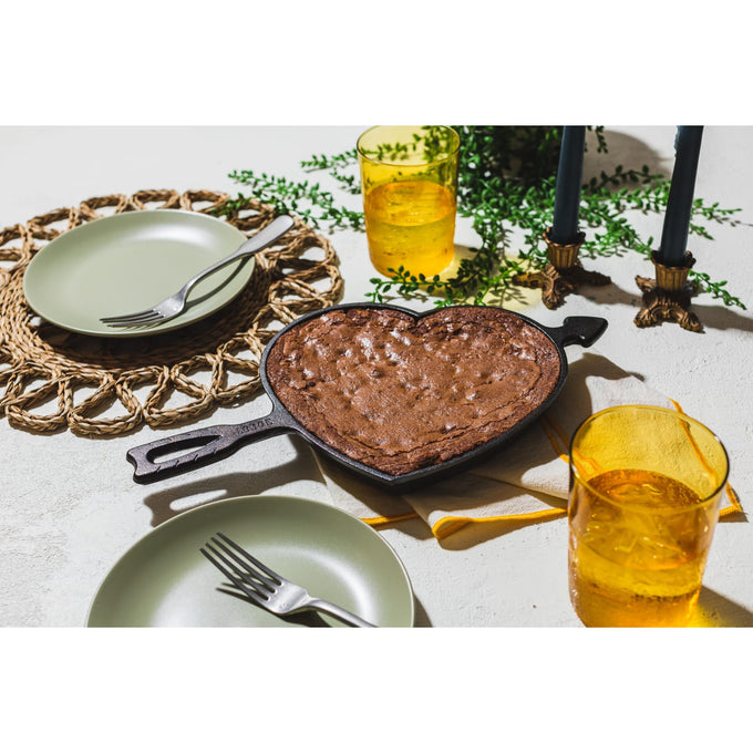 Handcrafted cast iron skillet in a heart shape with arrow handle, made in USA, featuring a freshly baked chocolate dessert, set on a table with green plates, yellow glasses, and decorative greenery, ideal for Kitchen & Food, Cookware, Cast Iron, Baking Supplies, USA Made, Best Sellers, and Promo Eligible collections.