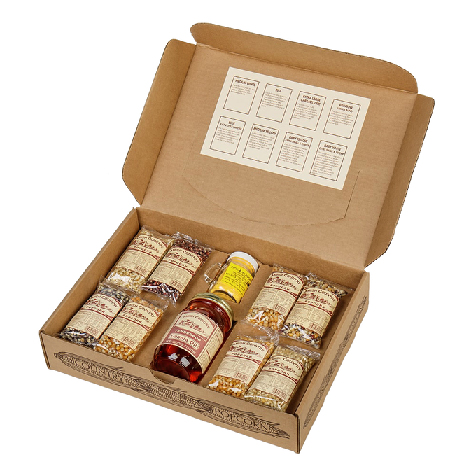 Open cardboard box containing eight small labeled bags of assorted handcrafted popcorn kernels, a bottle of Amish-made seasoning and a jar of syrup, suitable for Kitchen & Food, Snacks and Sweets, USA Made, Amish Made, and Best Sellers collections.
