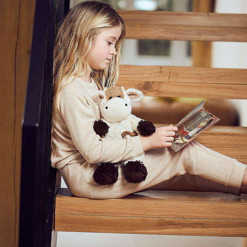 Young girl sitting on wooden stairs reading a book while holding a soft, weighted plush animal infused with natural lavender, part of health and wellness and children's toys collections.