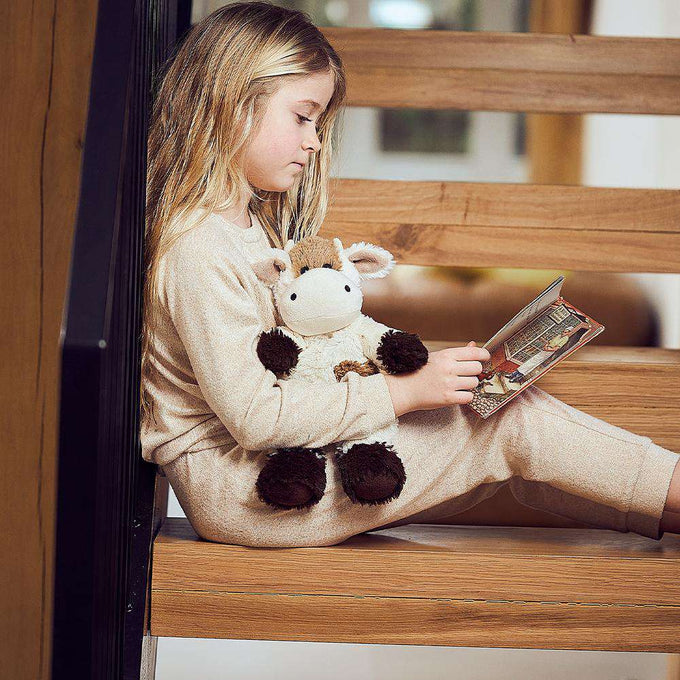 Young girl sitting on wooden stairs reading a book while holding a soft, weighted plush animal infused with natural lavender, part of health and wellness and children's toys collections.