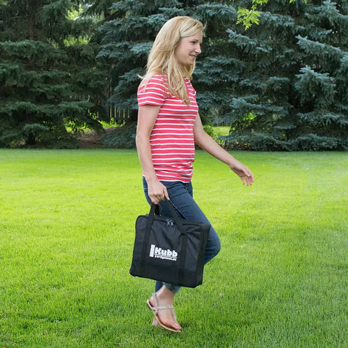 Woman carrying a black canvas bag with a logo, designed for easy transport of a handcrafted wooden outdoor lawn game set, pictured on a grassy lawn with trees in the background.
