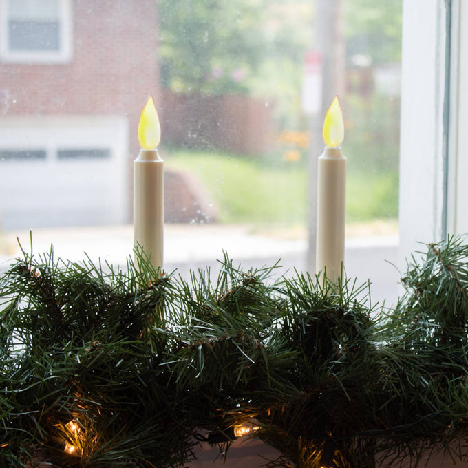 Pair of slim cordless LED taper candles with realistic amber flickering flames, nestled in green holiday garland on a windowsill, ideal for festive home decor collections.