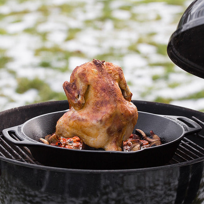 Golden whole chicken roasting upright on a cast iron poultry roaster with mushrooms and carrots in a cast iron pan on an outdoor grill, ideal for Kitchen & Food, Cookware, Cast Iron, Pots and Pans, Best Sellers, and Promo Eligible collections.