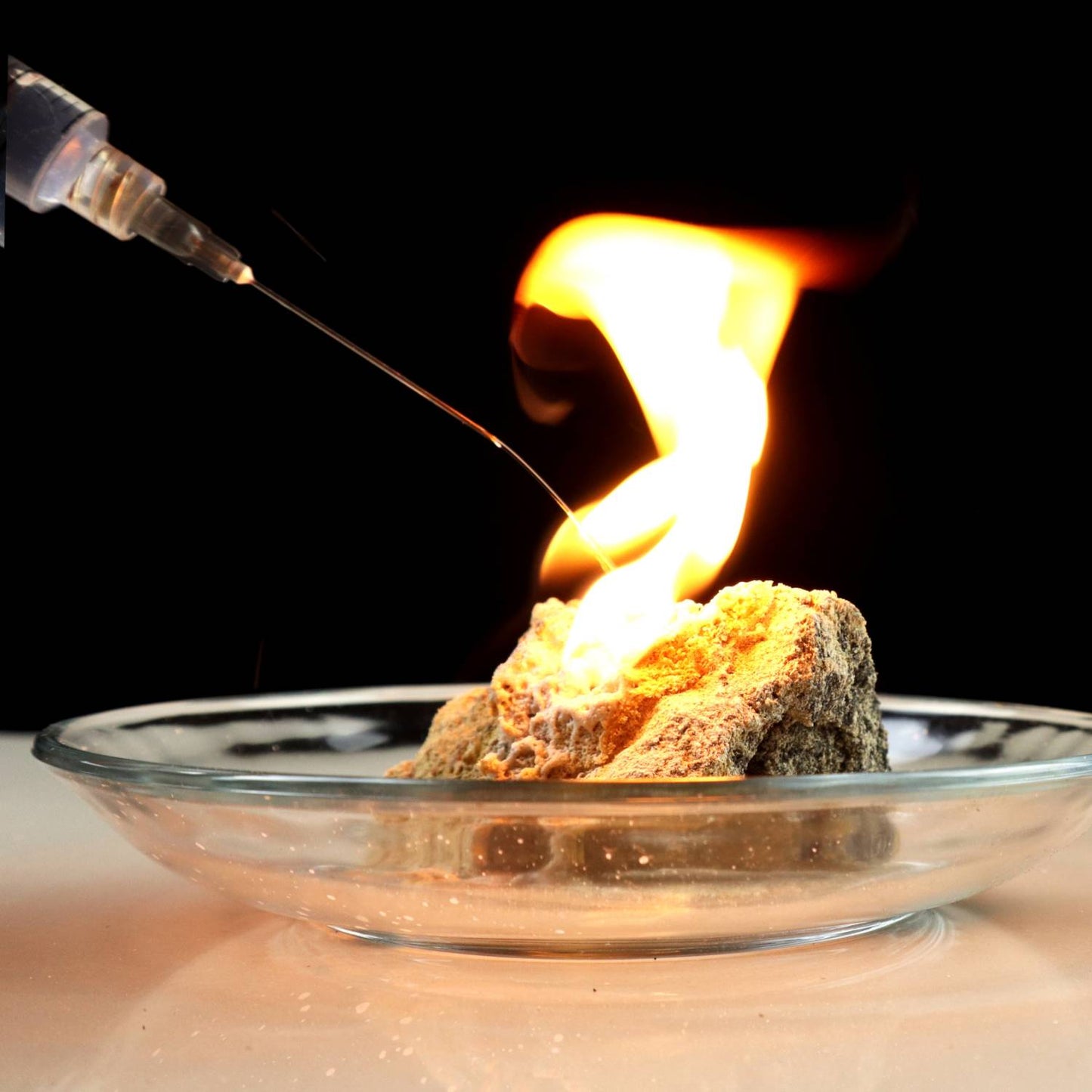 Close-up of a glowing chunk of miner's grade calcium carbide ignited with a flame, placed on a clear glass dish for demonstration, related to Lamps & Lighting and Lamp and Lantern Fuel collections.