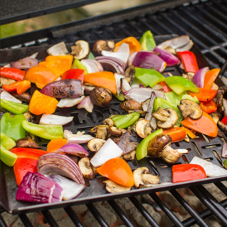 Seasoned steel grilling pan with an open grid design cooking colorful bell peppers, red onions, and mushrooms over a campfire grill in outdoor farm and camping setting.