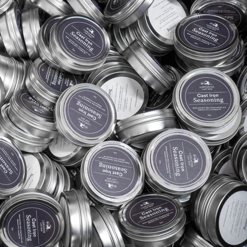 Multiple small metal tins with dark blue labels stacked together, containing handcrafted cast iron seasoning made in the USA, featured in Kitchen & Food, Cookware, and Cast Iron collections.