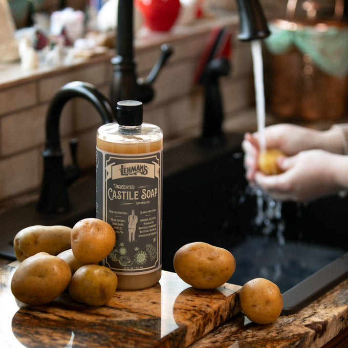 Amber bottle of Lehman's unscented castile soap on a kitchen countertop surrounded by potatoes, with a person washing potatoes under running water at the sink, highlighting handcrafted USA-made soap for Health & Wellness, Grooming and Personal Hygiene, Soaps and Soapmaking, and Best Sellers collections.