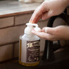 Hand dispensing natural foaming hand soap from clear bottle with pump top, featuring moisturizing formula with olive, coconut, honey, and jojoba oils in a Health & Wellness and Grooming collection.