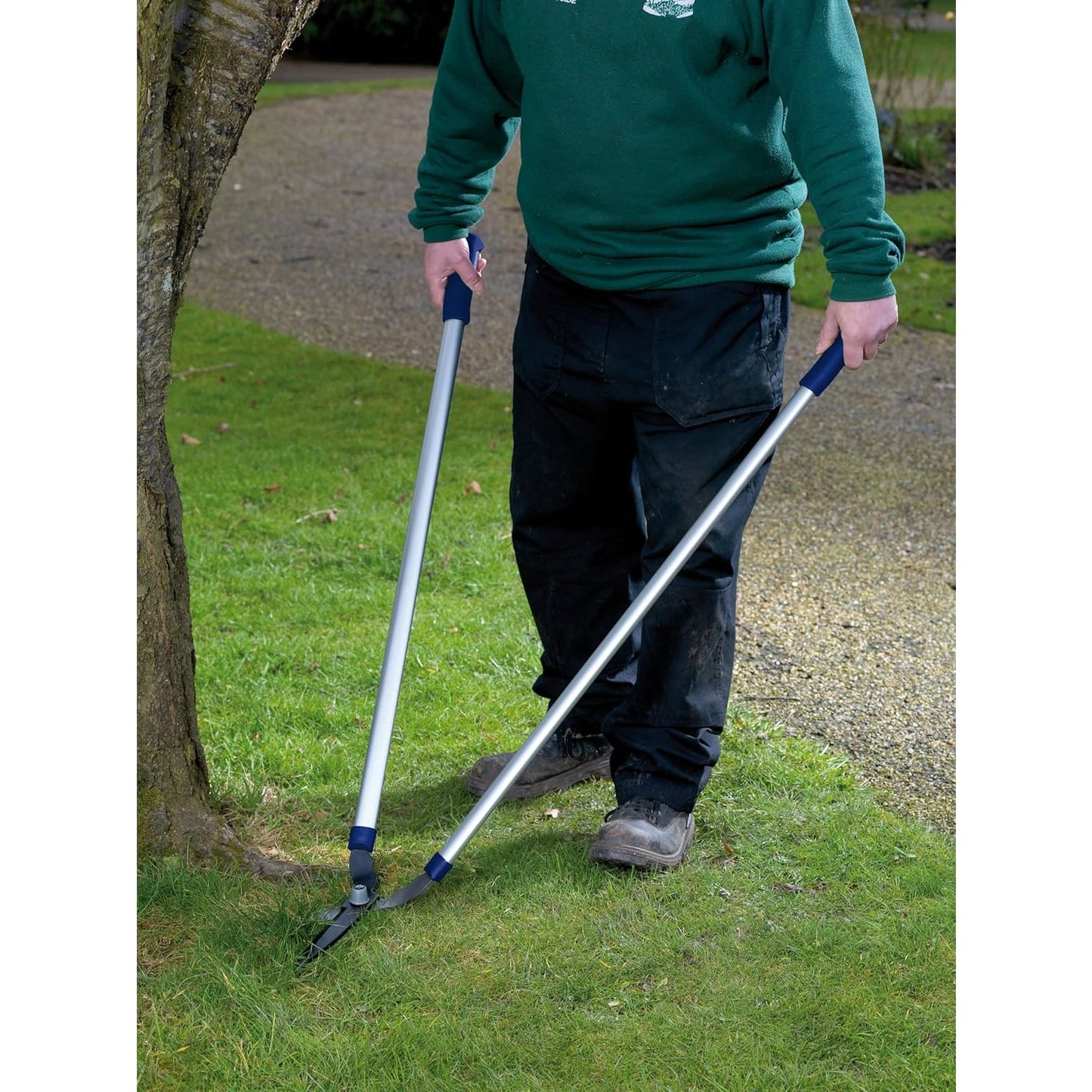 Person using long-handled garden shears with rust-resistant, tempered carbon steel blades to trim grass near a tree, suitable for farm garden and lawncare tasks.