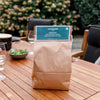 Brown paper bag of charcoal labeled for tabletop grill use, placed on a wooden outdoor dining table set with plates, glasses, and greenery in the background, ideal for grilling and stoves collections.