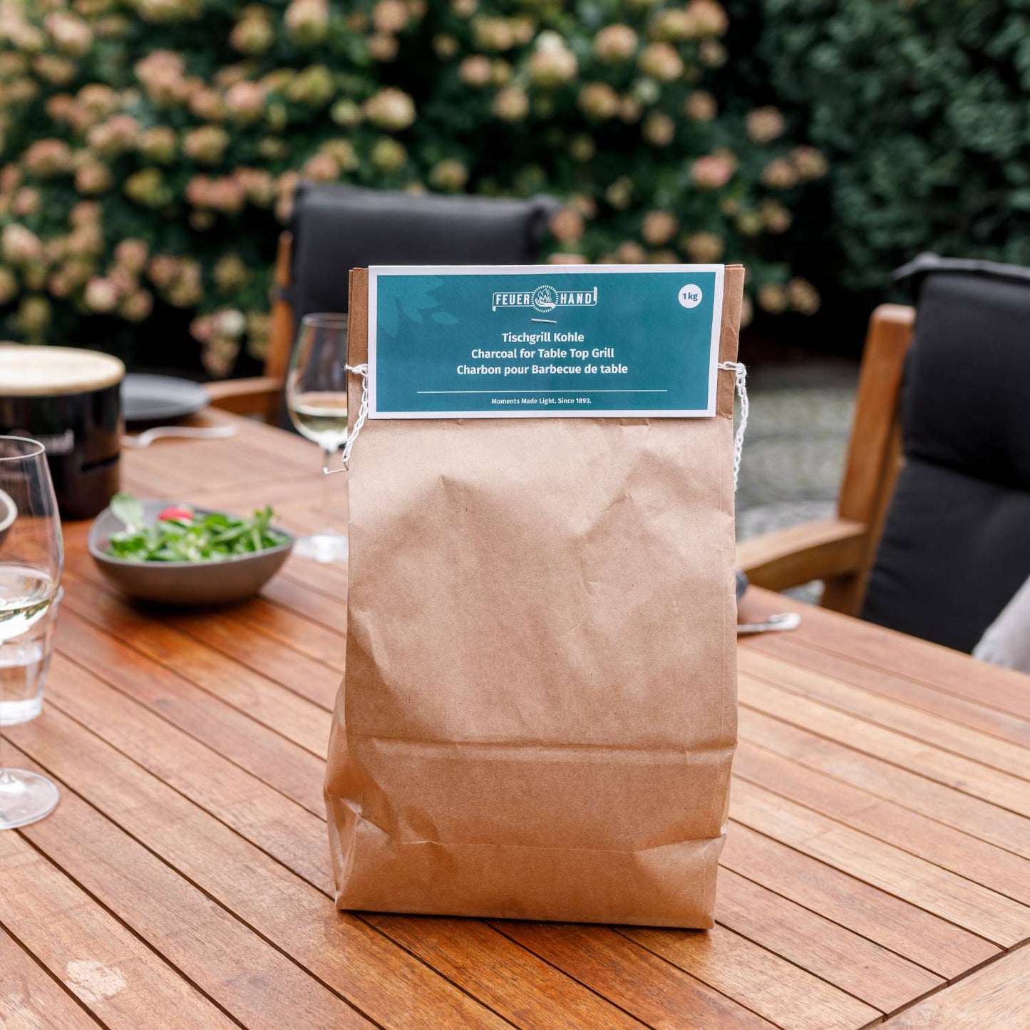 Brown paper bag of charcoal labeled for tabletop grill use, placed on a wooden outdoor dining table set with plates, glasses, and greenery in the background, ideal for grilling and stoves collections.