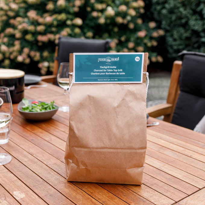 Brown paper bag of charcoal labeled for tabletop grill use, placed on a wooden outdoor dining table set with plates, glasses, and greenery in the background, ideal for grilling and stoves collections.