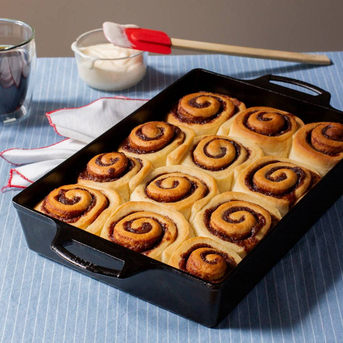 Handcrafted cast iron pan with dual handles baking a batch of golden cinnamon rolls, set on a blue striped cloth with a brush and bowl of icing, featured in Kitchen & Food, Cookware, Cast Iron, USA Made, Best Sellers, and Promo Eligible collections.