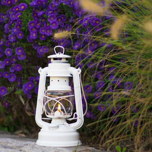 Handcrafted white galvanized oil lantern with thermal heat-resistant globe softly glowing on a stone surface surrounded by vibrant purple flowers and ornamental grasses, perfect for Camping, Lamps & Lighting, and Emergency Supplies collections.