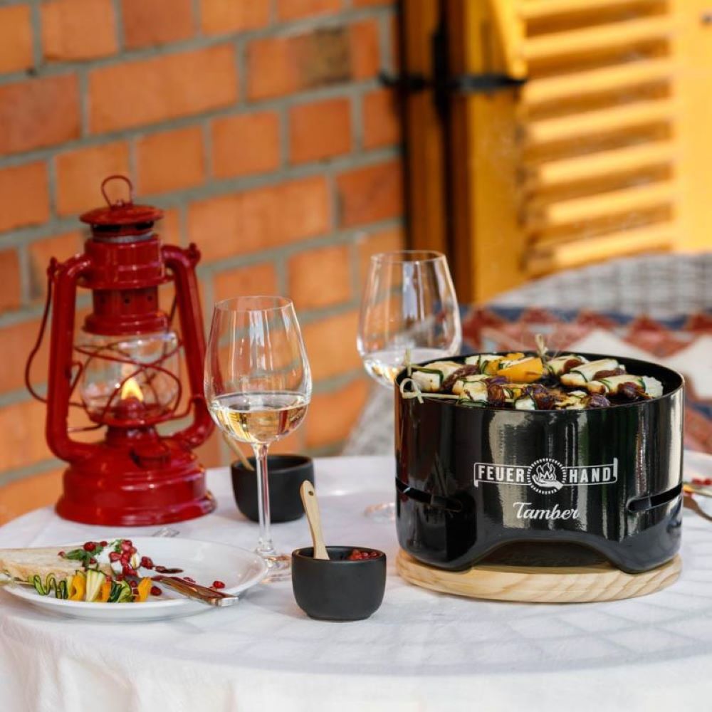 Glossy black mini charcoal grill with skewers cooking on a wooden table alongside wine glasses, a red lantern, and small dishes, perfect for cozy outdoor dining in Farm, Garden & Tool and Grilling collections.