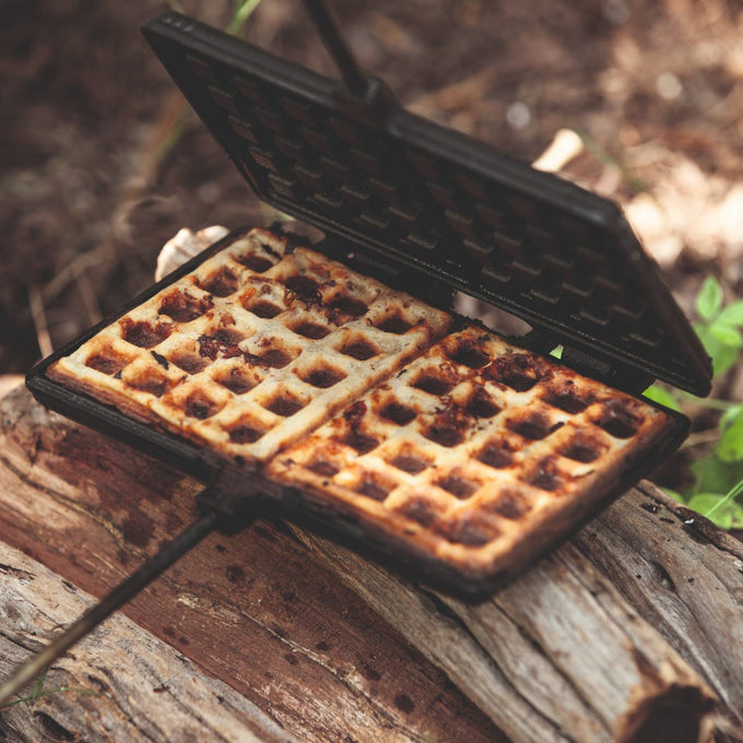 Cast iron waffle iron with a long handle cooking golden brown waffles over a campfire, perfect for outdoor adventures in Farm, Garden & Tool, Camping, Best Sellers, and Promo Eligible collections.