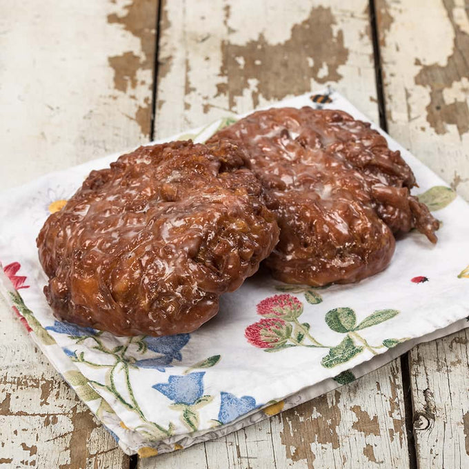 Two handcrafted glazed fruit fritters with a shiny sweet coating, resting on a floral cloth over rustic wooden surface, ideal for Kitchen & Food, Snacks and Sweets, Holiday Hosting, USA Made, Best Sellers, and Promo Eligible collections.