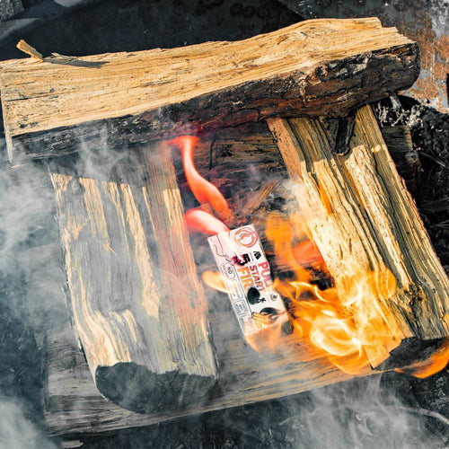 Fire starting tool igniting beneath stacked split wood logs with bright flames and smoke in outdoor camping or farm fire setting.