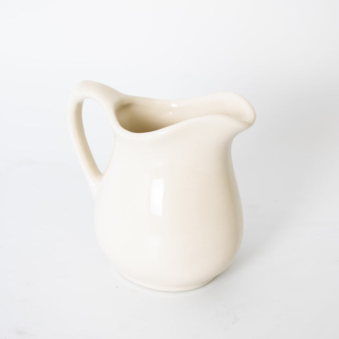 Cream-colored stoneware pitcher with a curved handle and rounded spout, handcrafted in the USA, ideal for serving in Kitchen & Food, Dinnerware, and Storage collections.