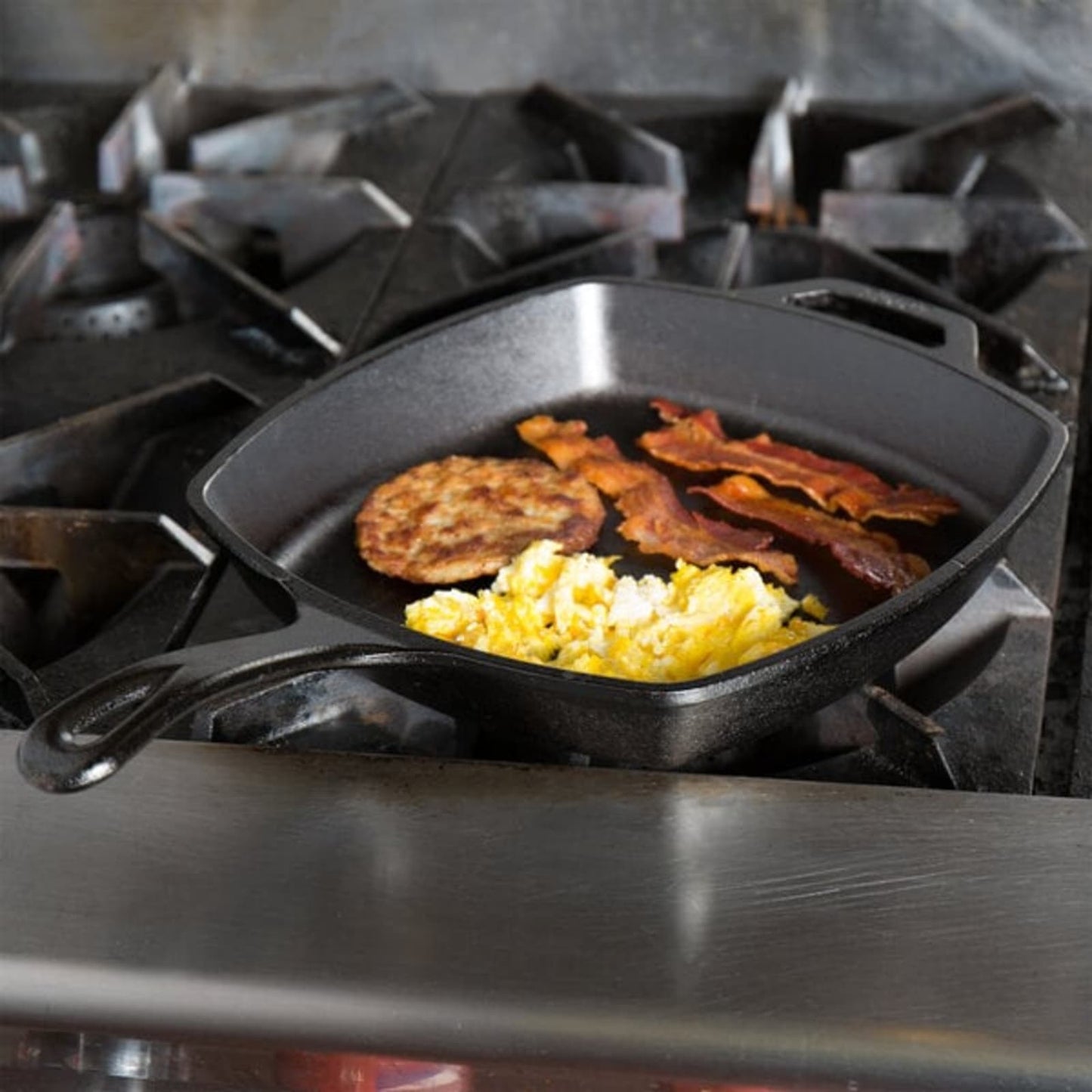 Square cast iron skillet cooking scrambled eggs, crispy bacon, and a sausage patty on a commercial gas stove, highlighting durable cookware for Kitchen & Food, Cast Iron, and USA Made collections.