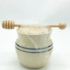 Handcrafted stoneware honey pot in rustic stone color with two classic blue stripes, covered with plastic wrap and topped with a wooden honey dipper, ideal for Kitchen & Food and Storage collections.