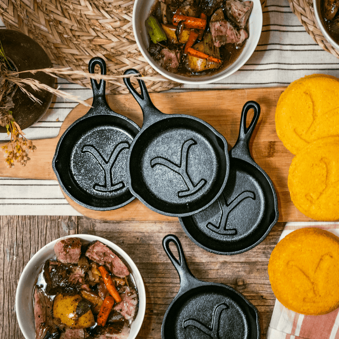 Set of handcrafted cast iron skillets with distinctive raised "Y" designs, arranged alongside bowls of hearty stew and golden cornbread, highlighting USA Made cookware for Kitchen & Food and Cast Iron collections.