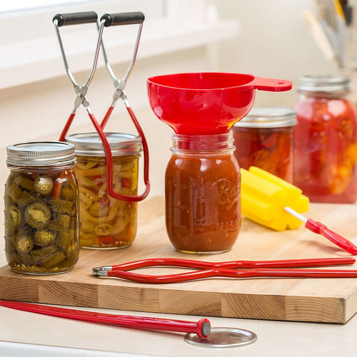 Set of durable plastic canning tools with bright red handles including jar lifter, funnel, and wrench, displayed with glass jars of preserved vegetables and sauces in a kitchen setting.