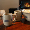 Two handcrafted stoneware mugs with blue stripe accents on a wooden table beside a matching bowl of popcorn, with a cozy fireplace in the background.