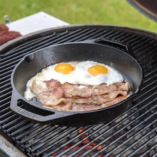 Handcrafted cast iron pan with short side handles cooking two sunny-side-up eggs and crispy bacon on an outdoor grill, ideal for Kitchen & Food, Cookware, Cast Iron, Best Sellers, and Promo Eligible collections.