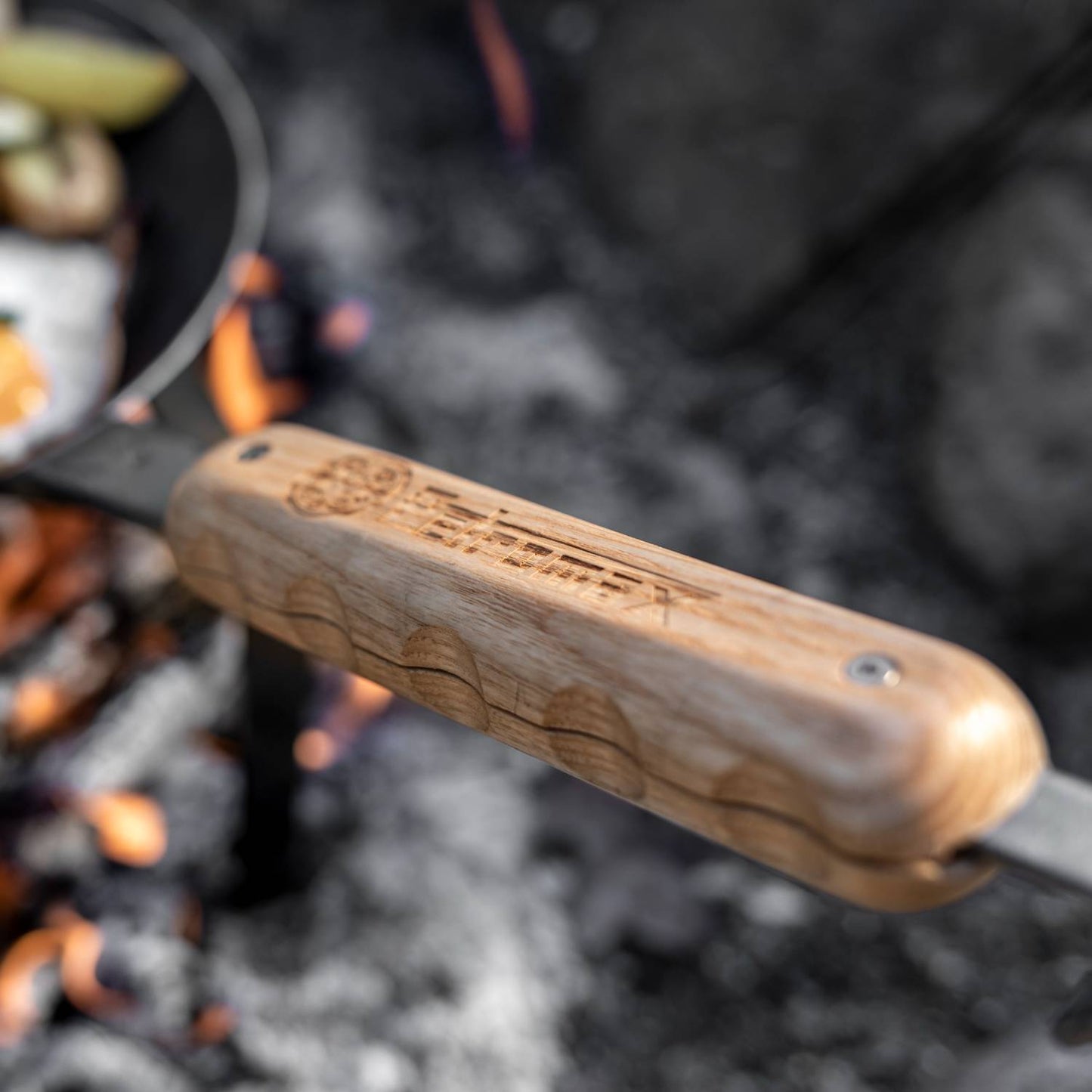 Close-up of a light wooden handle with ergonomic grooves and engraved logo, attached to a metal frame over glowing campfire coals, perfect for camping and cookware collections.