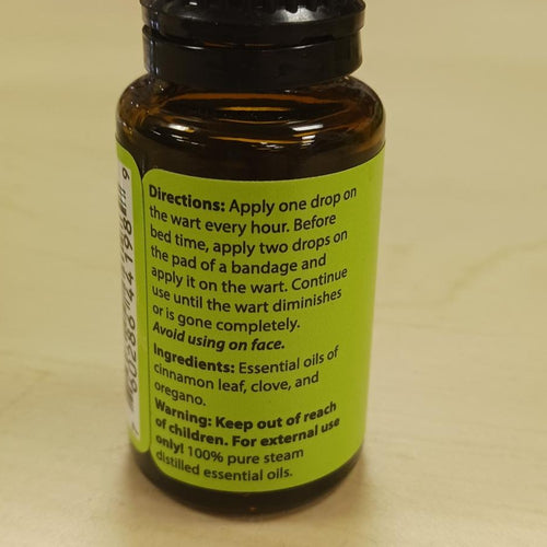 Amber glass bottle of all-natural wart oil featuring a green label with usage directions and ingredients, highlighting cinnamon leaf, clove, and oregano essential oils, made in USA.