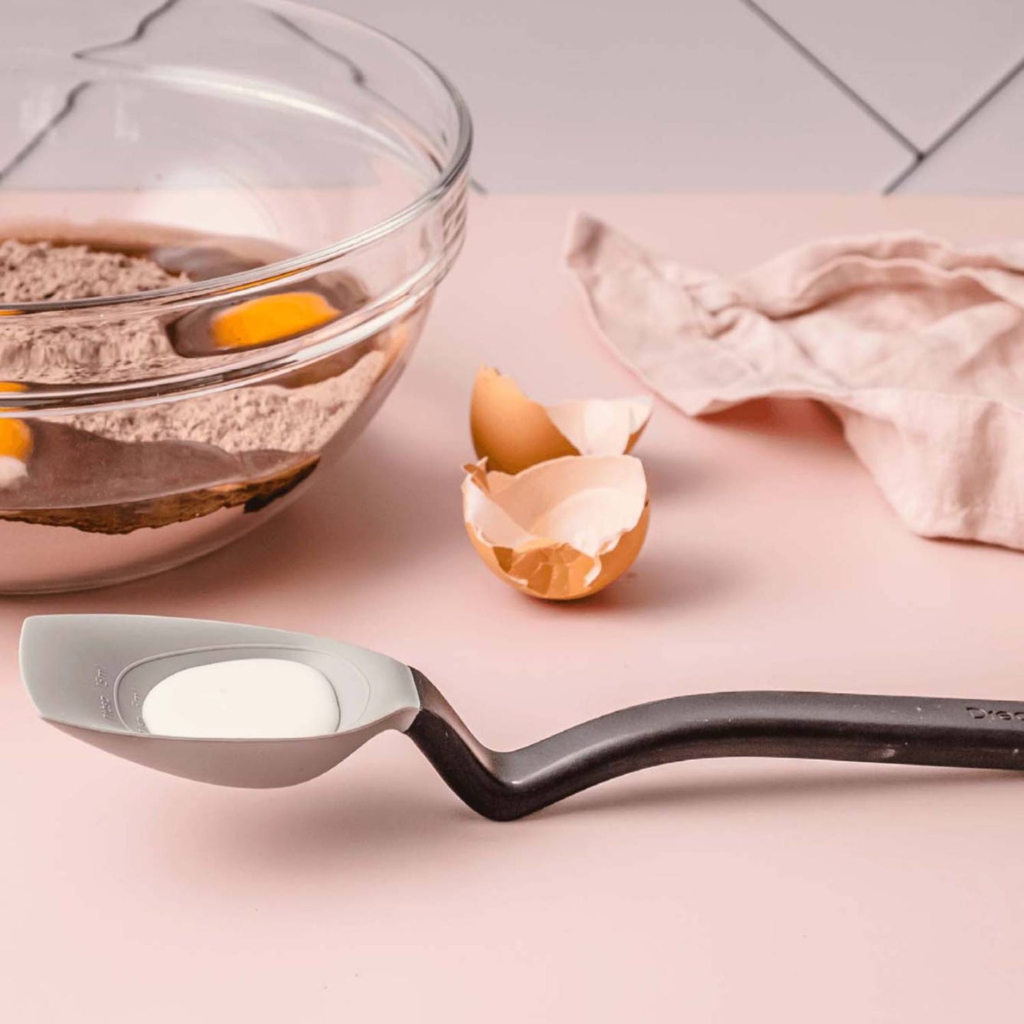 Flexible black-handled silicone spoon scooping liquid ingredients from a glass mixing bowl with cracked eggshells nearby, suited for Kitchen & Food, Food Prep, Measuring, and Cooking Helpers collections.