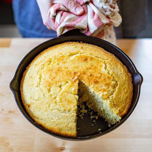 Freshly baked golden cornbread with a slice removed, held by hands using a cloth, in a pre-seasoned cast iron skillet designed for unmatched heat retention, featured in Kitchen & Food, Cookware, Cast Iron, Best Sellers, and Promo Eligible collections.