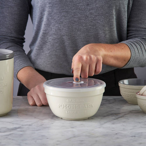 Hand pressing the airtight lid on a cream-colored Mason Cash all-purpose bowl with embossed pudding basin text, styled for Kitchen & Food, Cookware, and Storage and Serving collections.