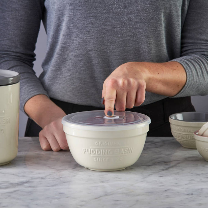 Hand pressing the airtight lid on a cream-colored Mason Cash all-purpose bowl with embossed pudding basin text, styled for Kitchen & Food, Cookware, and Storage and Serving collections.