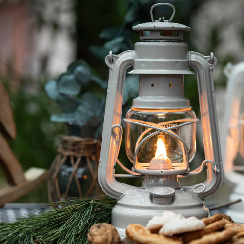 Handcrafted galvanized oil lantern with thermal heat-resistant globe glowing warmly outdoors on a rustic table with pine branches and natural decor, ideal for Lamps & Lighting, Camping, and Emergency Supplies collections.