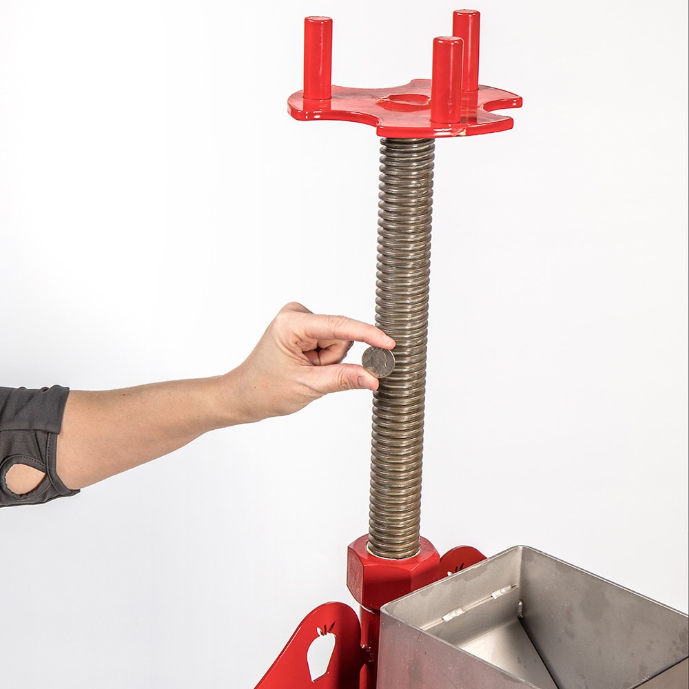 Close-up of a hand turning the threaded screw mechanism on a handcrafted all-stainless steel cider press with a red frame, designed for orchard and garden use in USA Made collections.
