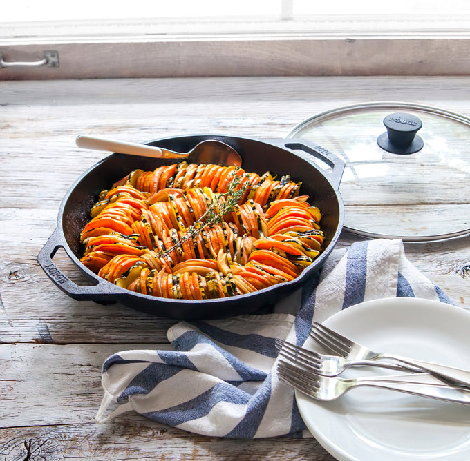 Handcrafted cast iron skillet filled with layered sliced vegetables seasoned with herbs, resting on a rustic wooden table with plates and forks for a cozy meal in Books and Cookbooks collections.