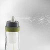 Clear glass oil spray bottle with a green silicone band and white spray nozzle releasing a fine mist, designed for precise oil application in kitchen and cookware use.